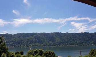 A view of a calm lake, surrounded by green hills and blue sky. Soft clouds drift across the landscape.