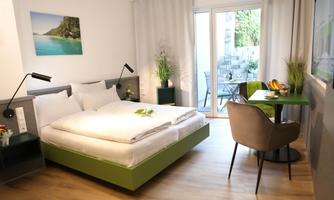 A modern room with a double bed and a small dining area. Large windows offer a view of a terrace with plants.
