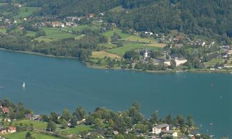 A picturesque landscape with a tranquil lake and surrounding mountains. In the background, houses and a church can be seen.