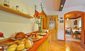 An inviting kitchen with a counter decorated with various baked goods, fruits, and a vase with flowers. In the background, tables and windows are visible, creating a cozy atmosphere.
