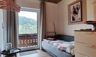 A cozy room with a single bed and a large window overlooking a green mountain landscape. A picture of a dog hangs on the wall, and there is a small desk.