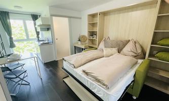 A modern bedroom with a large bed and wooden shelves. The room has large windows and is furnished with simple furniture.