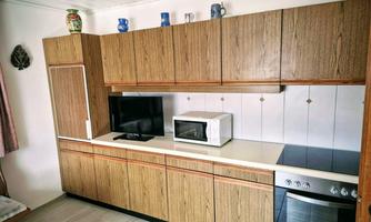 A modern kitchen with wooden furniture and white tiles. Featured are a television, a microwave, and a stove.