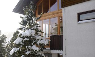 A modern house in the snow with large windows and a balcony. Surrounded by snow-covered trees and a wintry landscape.