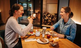 Two women are sitting at a table, enjoying a breakfast with various dishes. In the background, a fireplace is burning, creating a cozy atmosphere.