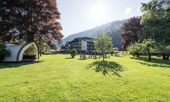 A spacious garden with green grass and beautiful trees. In the background, a charming hotel is visible against a picturesque mountain landscape.