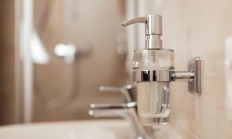 An elegant soap dispenser made of glass with a shiny metal pump mechanism. In the background, a modern bathroom with soft color tones can be seen.