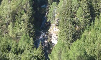 A picturesque waterfall flows through dense, green forests. Sun rays illuminate the surroundings and create a peaceful atmosphere.