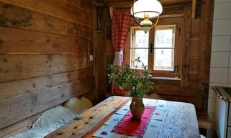 Cozy dining area in a wooden cabin with an old table and a vase full of flowers. A window with red curtains lets in plenty of light.