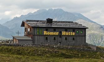 An alpine hut named "Römer Hütte" stands in a natural mountain landscape. In the background, high mountains and gray clouds can be seen.