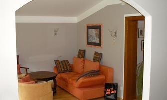 A cozy living room with an orange sofa and a round table. The walls are painted in light gray tones, and there are decorative pictures as well as a floor lamp.