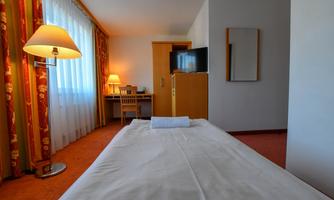 A cozy hotel room with a single bed, a desk corner, and a television. The room is bright with windows and curtains in warm colors.