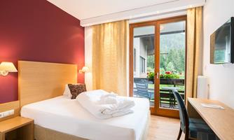 A cozy hotel room with a large bed and natural light. The windows offer a view of a balcony with flowers.