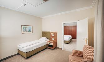 A simple hotel room with a single bed, a nightstand, and an armchair. There is a wardrobe and access to another room.