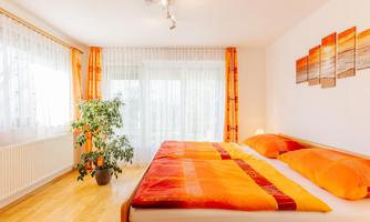 A bright bedroom with a large double bed and decorative orange pillows. Large windows provide plenty of daylight and a cozy atmosphere.