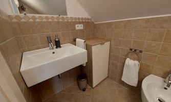 A modern bathroom with a simple sink installation and earth-toned tiles. The furnishings include a small refrigerator and a bidet.