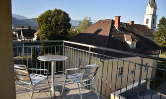 A balcony with a small table and two chairs. In the background, houses and a church can be seen, surrounded by trees and mountains.