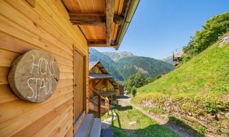 A rustic wooden house with a hand-painted sign. In the background, green hills and mountains can be seen under a clear blue sky.