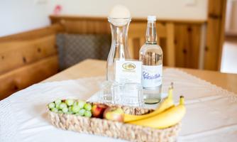 A table with a glass water bottle, a carafe, and two glasses. Next to them lies a basket with apples, bananas, and grapes.