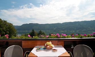 A beautiful view from a terrace of a lake and the surrounding mountains. On the table, there are flowers and a fruity decoration visible.