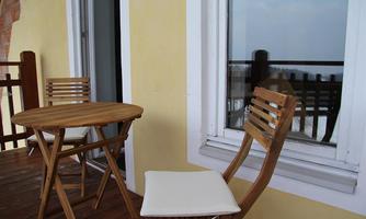 A small balcony with a round wooden table and two woven chairs. The wall is painted in a soft yellow and there is a window with a view.