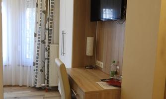 A narrow room with a desk, a TV on the wall, and a window with curtains. The floor is made of wood and the walls are brightly designed.