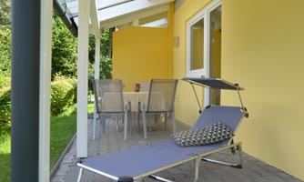 A cozy terrace with a lounger and a table with chairs. The walls are painted in a warm yellow shade, surrounded by beautiful green plants.