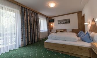 A cozy bedroom with a large bed and warm wooden decor. Large windows provide plenty of daylight and a view of the outside.