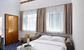 A modern bedroom with a bed and a cozy sofa. Large windows provide plenty of daylight and a warm atmosphere.