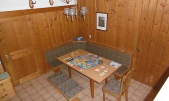 A cozy room with wooden walls and a table prepared for a board game. There is a seating area with cushions and ceiling lighting.