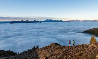 Two hikers are walking over a mountain with clouds that look like a sea. In the background, majestic mountains are visible.