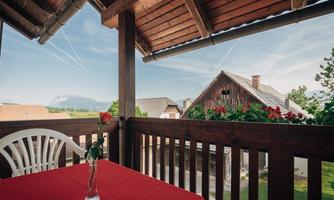 A balcony with a red table and a rose in a vase. In the background, wooden buildings and a clear sky are visible.