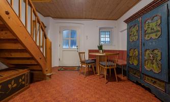 A cozy room with a wooden staircase, a round table, and chairs. In the background, there is a decorated cabinet and light is falling through a window.