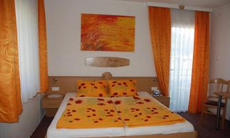 A cozy bedroom with a double bed and orange bedding. The walls are decorated with orange curtains and a colorful painting.