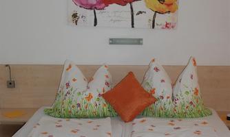 A bright bedroom with a double bed decorated with colorful pillows and blankets. A painting of flowers hangs on the wall.
