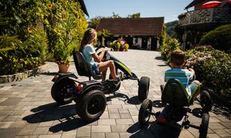 Two children are riding on go-karts on a paved path. In the background, plants and buildings can be seen.