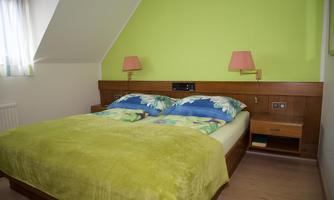 A cozy bedroom with a double bed and colorful bedding. The walls are painted in a fresh green hue, and there are two bedside lamps.
