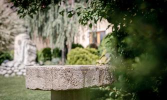 A round stone table stands in a green garden. In the background, trees and a decorative sculpture are visible.