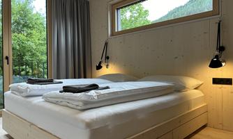 A cozy bedroom with a large double bed and wooden details. Daylight shines through the windows and there is a view of the greenery.
