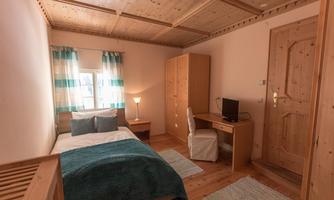 A simple bedroom with a single bed, a desk, and a chair. The walls are light and the room has a cozy atmosphere.