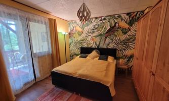 A cozy bedroom with a large bed and a colorful wall decoration made of tropical plants. A pleasant atmosphere is created by the natural light and the wooden furniture.