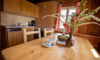A cozy kitchen with light wooden furniture and a large table. On the table sits a vase with fresh flowers and a cup.