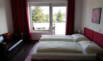 A cozy bedroom with two beds and a black sofa. Large windows offer a view of the nature outside.