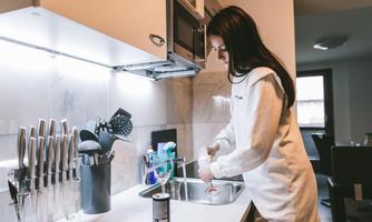 A person is washing dishes in the sink in a modern kitchen. In the background, various kitchen utensils and cleaning products are visible.