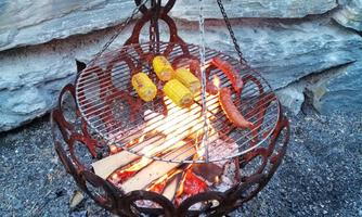 A grill with sausages and corn cobs is sizzling over an open fire. The setting is rustic with stones in the background.
