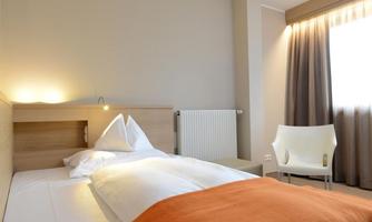 A modern bedroom with a double bed and bright decor. A chair and a window with curtains complete the room.