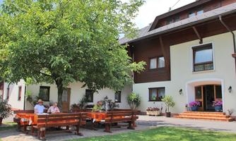 An idyllic house with a large tree in the garden. Two people are sitting at a table and enjoying the sun.