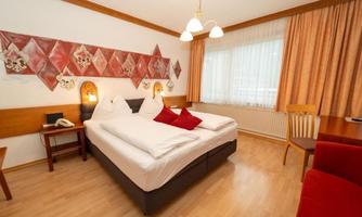 A cozy hotel room with a double bed and a red decorative pillow. Large windows provide plenty of daylight and an inviting atmosphere.