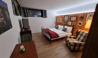 A cozy bedroom with a double bed, an armchair, and a small table. The walls are decorated with pictures and wood paneling.