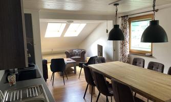 A bright living room with a large dining table and comfortable chairs. In the background, there are windows with natural light and a cozy sitting area.